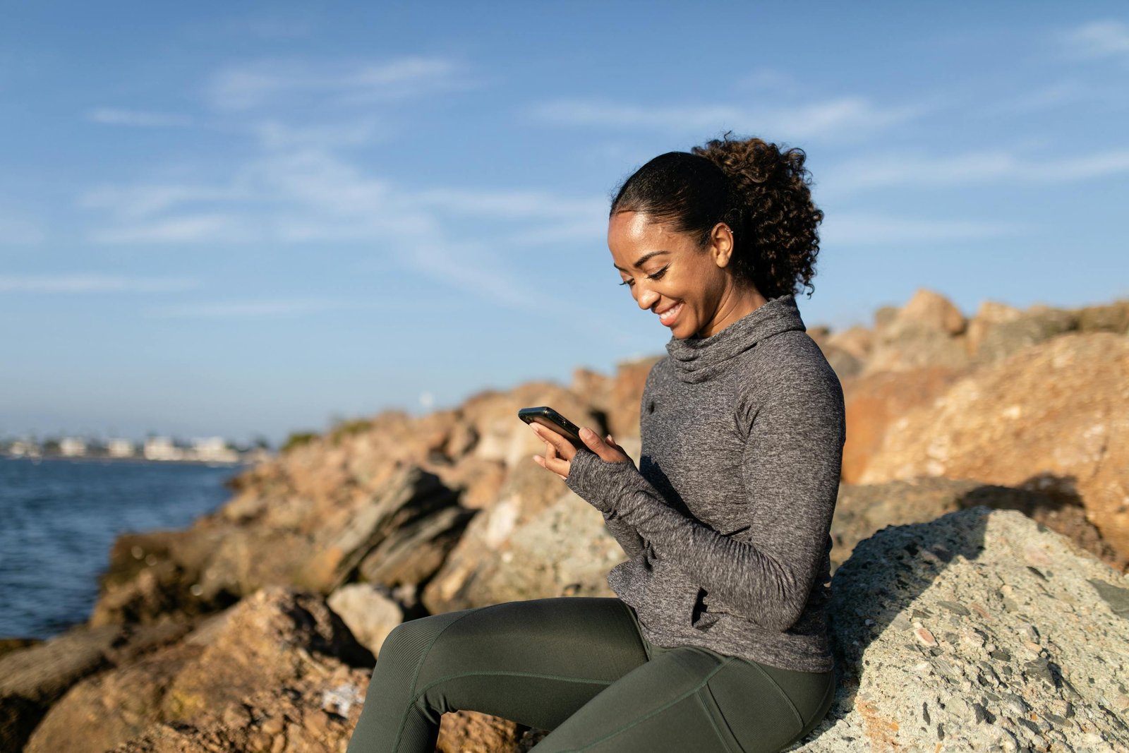Smiling woman using smartphone on rocky shore, enjoying a sunny day.